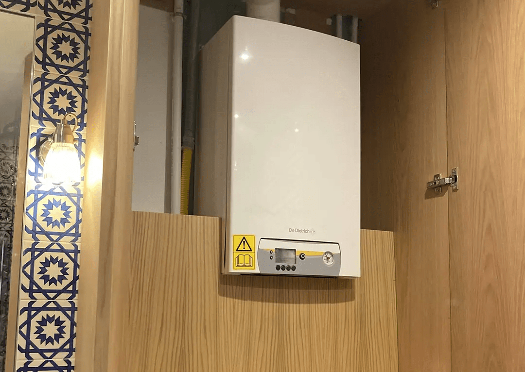 un chauffe-eau ou une chaudière encastrée dans un meuble en bois. Les murs sont décorés de carreaux à motifs bleus et blancs, ajoutant une touche esthétique.